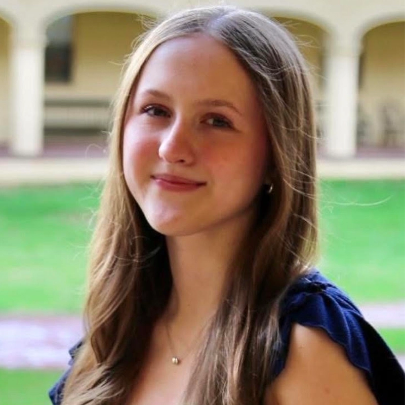A young woman with long, light brown hair smiles softly outdoors. She wears a navy dress and a small necklace. In the blurred background are green grass and arched beige columns. The lighting is natural, capturing the quiet elegance of Logan Altenburger in this serene setting.