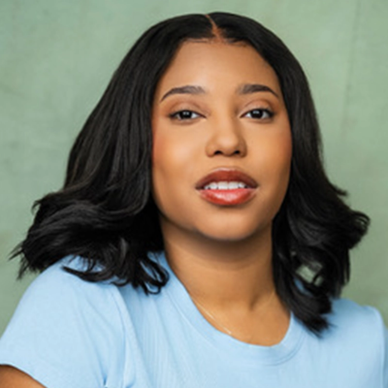 Alyssa Enita Stanford, a woman with medium skin, shoulder-length wavy black hair, and wearing a pale blue T-shirt, poses in front of a muted green background. She looks toward the camera with a slight smile, lips parted, and a confident, relaxed expression.
