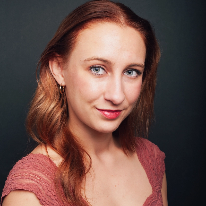 A woman with long, wavy auburn hair and blue eyes looks at the camera, smiling slightly. She wears a rose-colored lace top and gold hoop earring. The background is dark and plain, making Carly Ameling stand out in the softly lit portrait.