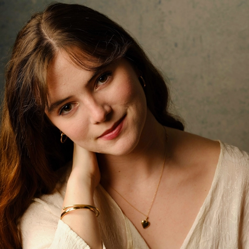 A young woman with long brown hair and fair skin tilts her head, gently touching her neck. Celeste Zwingel wears a cream-colored blouse, a gold heart necklace, hoop earrings, and a gold bracelet. She smiles softly against a neutral, textured background.