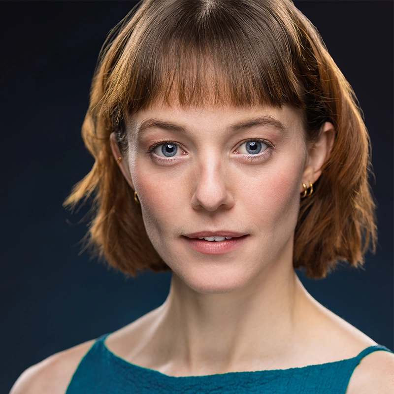 A woman with short, light brown hair and bangs is facing forward, wearing gold hoop earrings and a teal sleeveless top. She has fair skin, blue eyes, a neutral expression, and is photographed against a dark gradient background—a portrait reminiscent of Kendall Tubbs’ poised elegance.