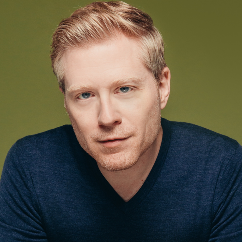 A man with short blond hair and light facial stubble wears a navy blue V-neck sweater. He has light skin, blue eyes, and a neutral expression. The background is a solid olive green color.