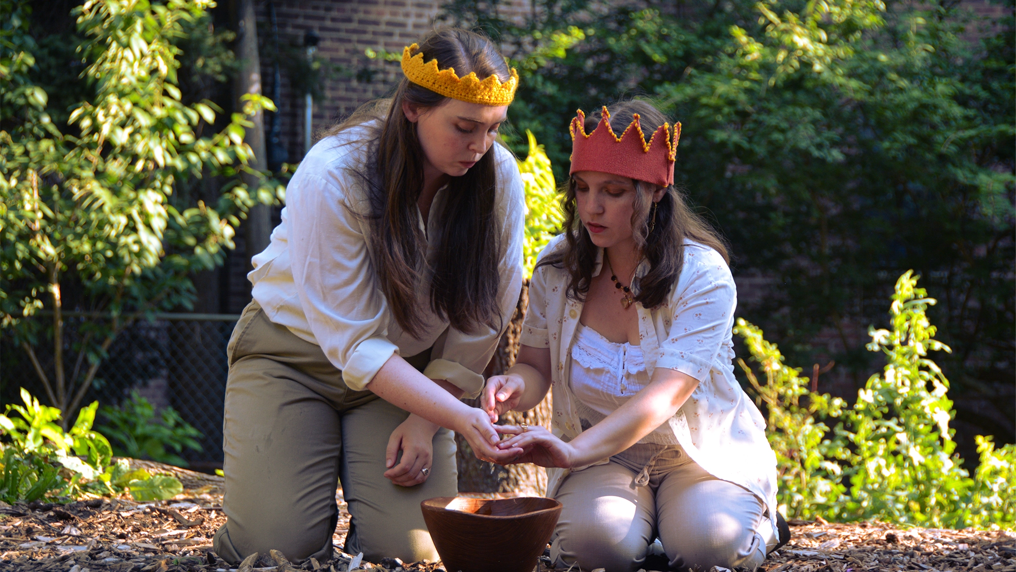 Anti-Album site image 26 Two women kneel outdoors in dappled sunlight, both wearing golden crowns as if presiding over their own private Anti-Album release concert. One pours something into the other’s hand, and they focus intently on the task, with a brown bowl on the ground in front of them. Lush green foliage and a fence are in the background.