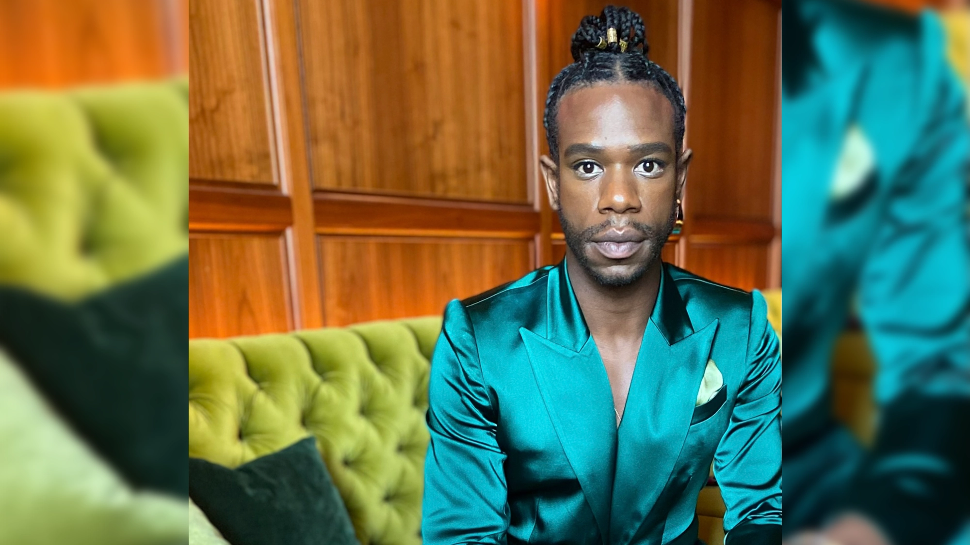 A person with braided hair in a top knot sits on a green tufted couch. They wear a silky teal blazer with a pocket square. The wooden paneled wall and warm lighting create an elegant, modern atmosphere, reminiscent of an intimate live listening session for Antwayn Hopper’s debut album. Their expression is serious and composed.