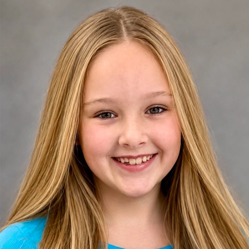 Smiling young girl with long straight blonde hair, wearing a light blue top. She is facing forward against a plain gray background. Her teeth are slightly visible, and her skin is fair. The lighting is even, and the portrait is clear and well-lit, capturing a look reminiscent of Kelsey Valente.