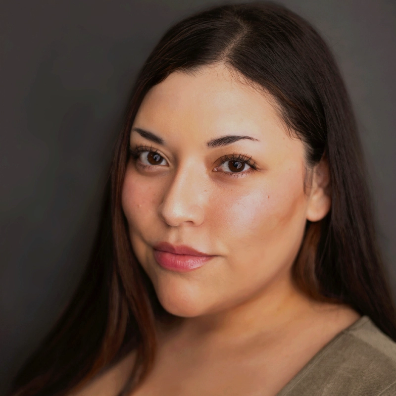 A woman with long, straight brown hair and light skin looks at the camera with a soft smile. She has brown eyes, subtle makeup, and is wearing a light olive top. The background is plain and dark, making her face the focal point. Her calm presence captures the quiet confidence of Samara Ariel.