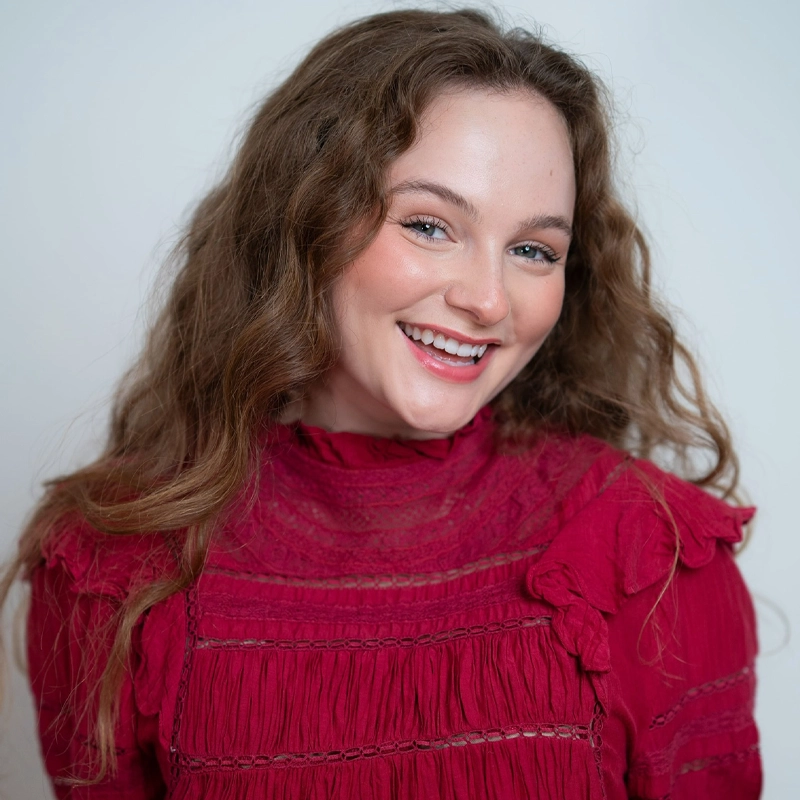 A smiling young woman—Isabel Asher—with long, wavy brown hair wearing a textured, bright magenta blouse with ruffled details stands against a plain light background. She looks directly at the camera, showing a joyful and friendly expression.