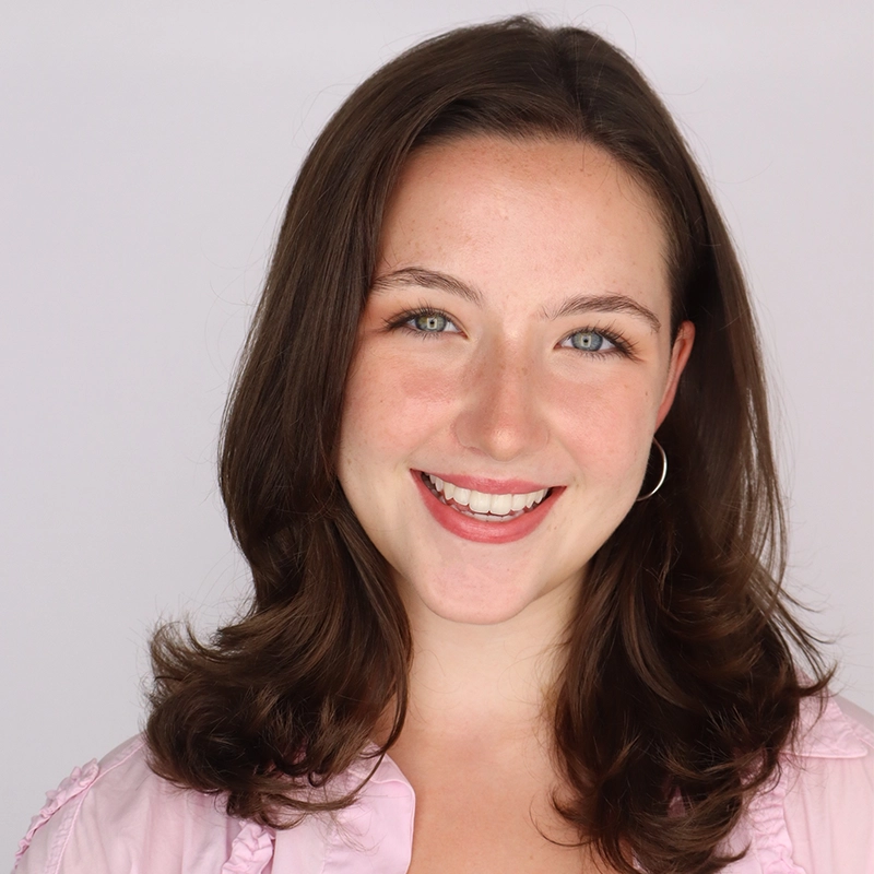 Young woman with fair skin, shoulder-length brown hair, and blue eyes smiles at the camera. She wears a pale pink blouse and silver hoop earrings, standing in front of a plain light background. Her expression is friendly and warm.