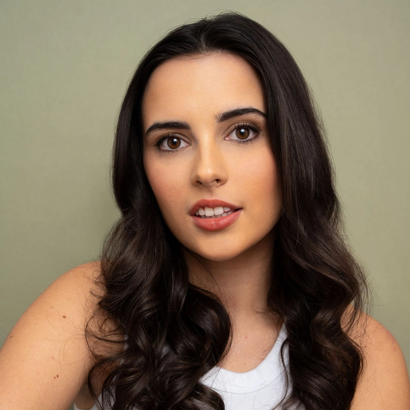 A young woman with long, dark, wavy hair and brown eyes looks into the camera. She wears a white sleeveless top and natural makeup. The background is a plain, light olive color. Her expression is neutral and relaxed.