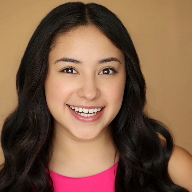A young woman with long, wavy dark hair smiles widely. Madeline Ayala has light brown skin, dark eyes, and is wearing a bright pink sleeveless top. The background is a solid tan color.