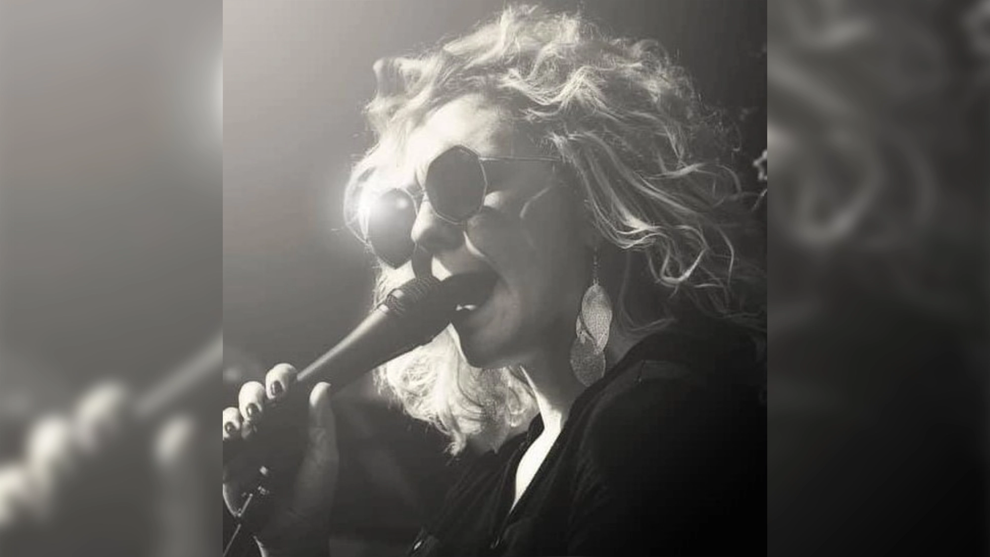 B.D. Greer site image 26 A person with curly hair and sunglasses sings into a microphone. Channeling the spirit of Jazz divas like B.D. Greer, they wear large, round earrings and dark clothing. Bright light shines from behind, creating a dramatic black-and-white portrait. The atmosphere suggests a live music performance.