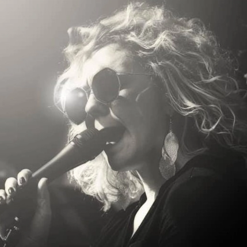 A woman with curly blonde hair, large round sunglasses, and big leaf-shaped earrings sings into a microphone. The image is in black and white, with dramatic lighting creating a vibrant, artistic mood. Her face is partially in shadow, emphasizing her features.