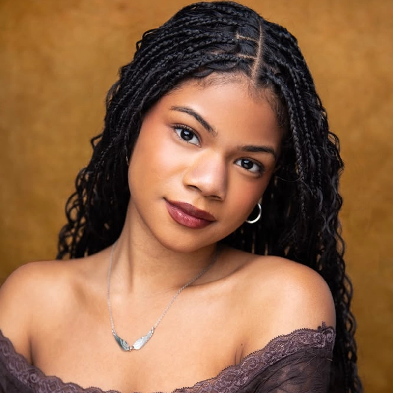 A young woman with long black braids, wearing a brown off-shoulder lace top, silver hoop earrings, and a necklace with a wing pendant. She has warm-toned skin, brown eyes, and dark lipstick. The background is a textured golden yellow. This striking portrait captures Keila Casasola Beaubrun’s unique style and radiant presence.