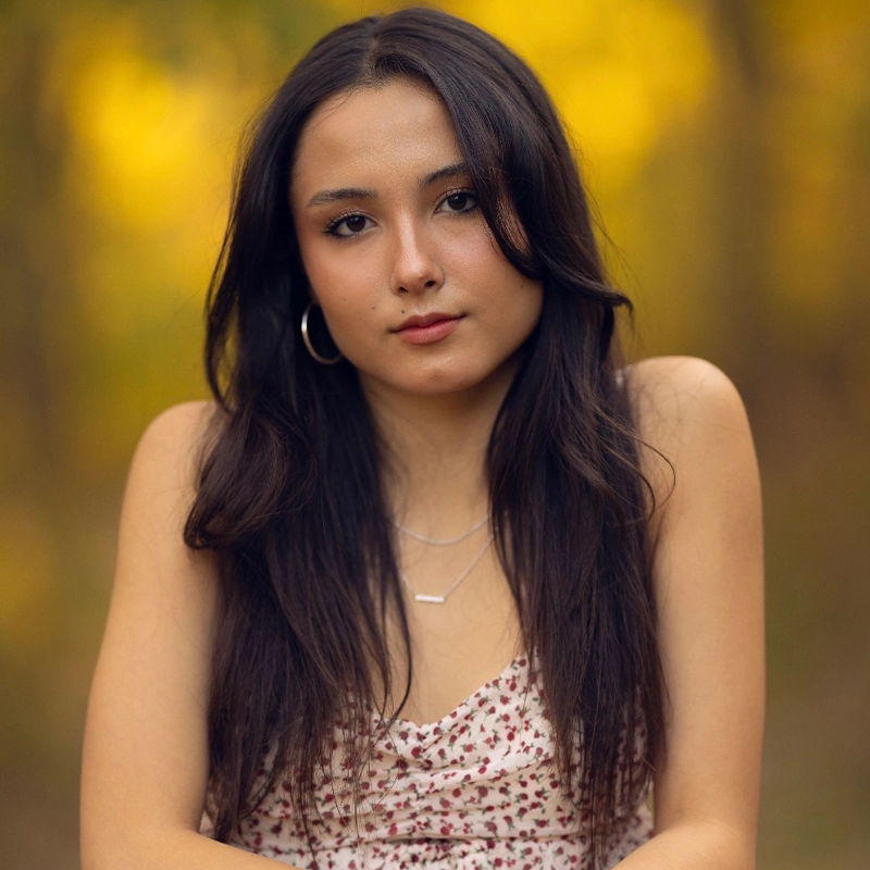 Ami Bellings, a young woman with long dark hair and hoop earrings, stands outdoors. She wears a white floral dress and layered necklaces. Her expression is neutral, and the blurred golden-yellow background suggests a natural, sunlit setting.