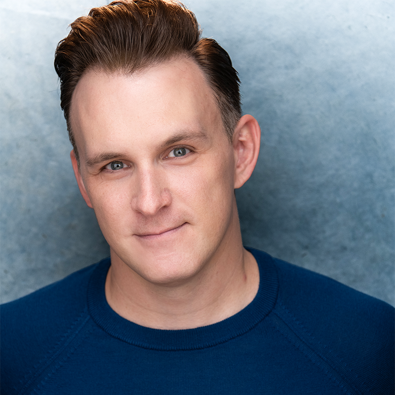 A man with fair skin and short, slicked-back brown hair wears a blue crew neck shirt, smiling slightly. The background is a soft, textured gray-blue. He is facing forward, well-lit, and the portrait is cropped at the shoulders.