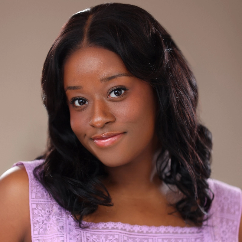 Jaylyn Tranice Bentley, a woman with medium-dark skin and wavy dark hair, smiles gently at the camera. She wears a lilac lace top and subtle makeup. The background is a soft, neutral color, making her the focal point of the image.