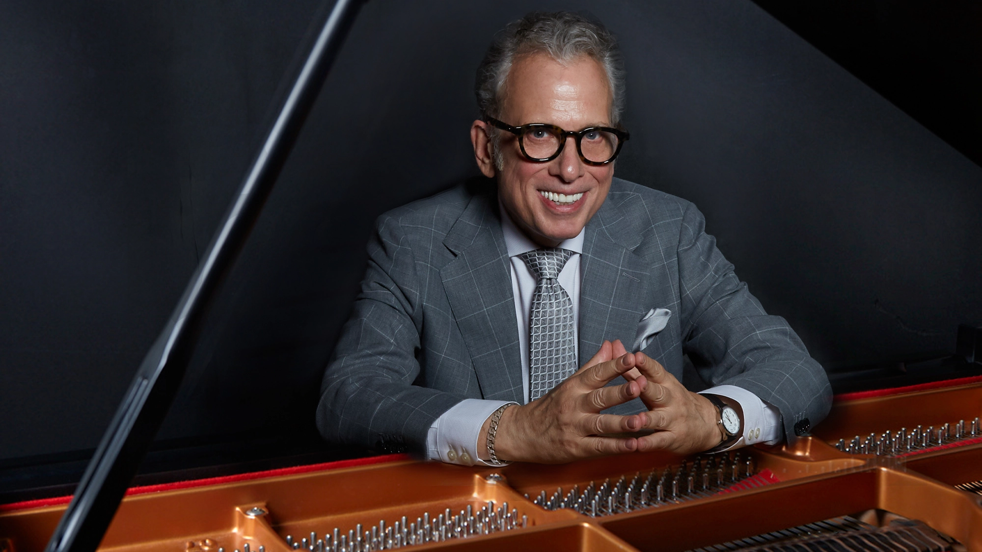 A smiling man in a gray plaid suit, patterned tie, and glasses sits at a grand piano, hands clasped. The piano’s strings and frame are visible in the foreground; the background is dark, highlighting the man and the instrument. Renowned pianist Billy Stritch pays tribute to Tony Bennett’s remarkable legacy—celebrating 100 Years of timeless music.