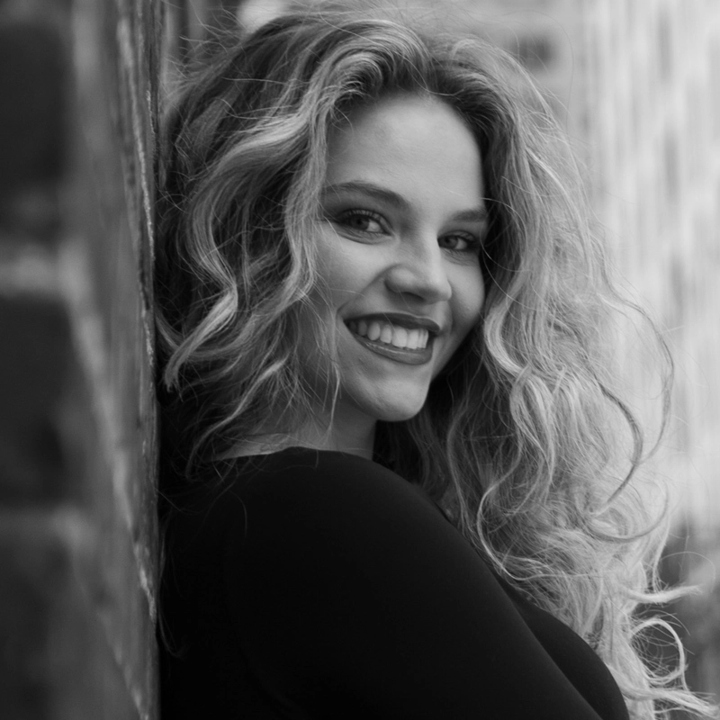 Black-and-white photo of a young woman, Eliza Boland, with long, wavy hair, smiling broadly. She wears a dark top and is leaning against a brick wall, with an urban background blurred behind her. The mood is cheerful and the lighting highlights her face.