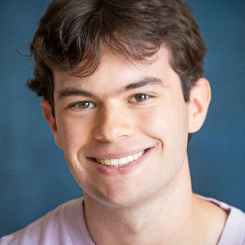 Billy Bollbach, a young man with short brown hair and fair skin, smiles at the camera. He is wearing a light pink shirt and is positioned against a soft blue background. The photo is bright, with even lighting highlighting his face.