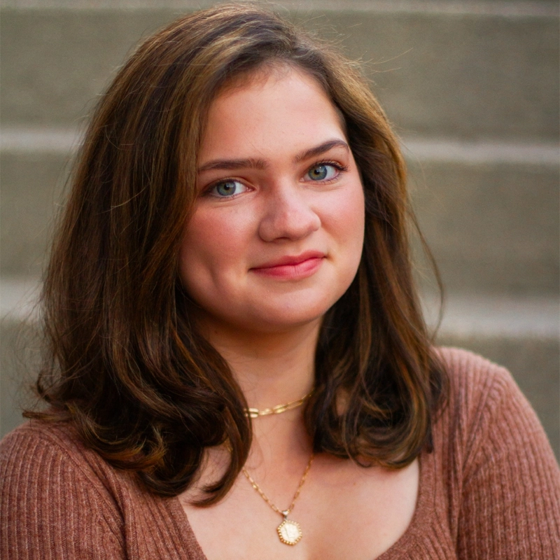 Liliana Borges, a young woman with shoulder-length brown hair and blue eyes, wearing a brown top and gold necklace, smiles softly at the camera. The background is blurred, showing neutral-colored steps.
