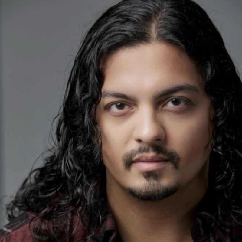 A man, identified as Dante Brattelli, with long, wavy dark hair and a trimmed goatee looks directly at the camera with a neutral expression. He is wearing a dark maroon shirt, and the background is softly blurred gray. His eyes are brown and his expression is calm.