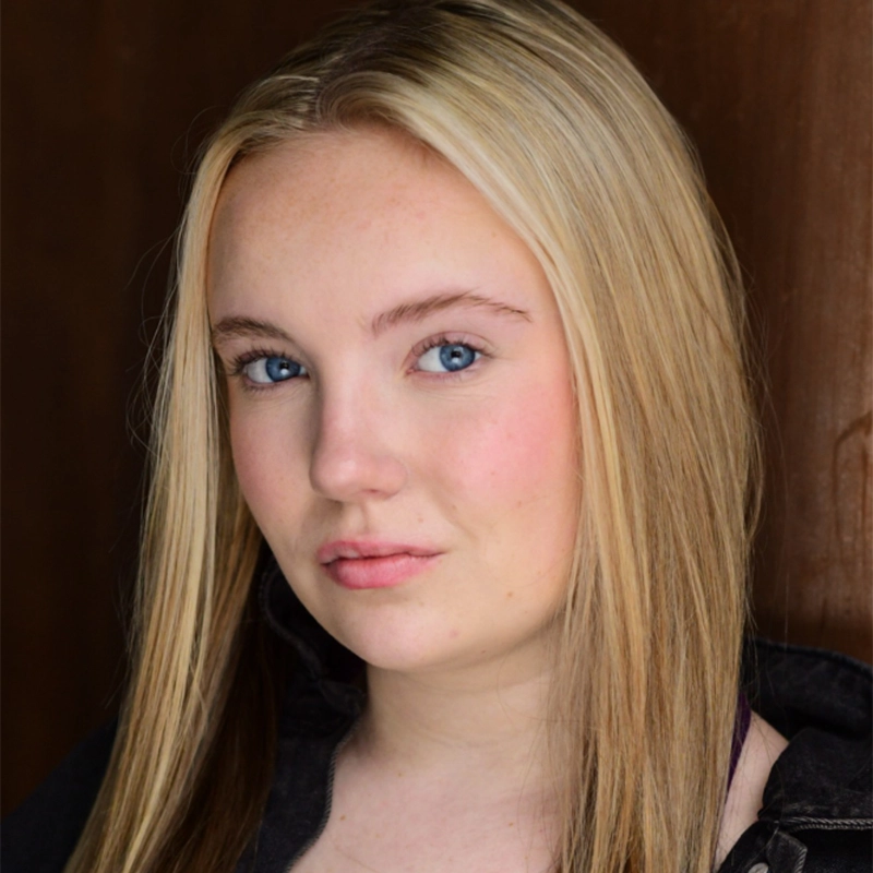 A young woman with long straight blonde hair and blue eyes looks directly at the camera. She has fair skin, light makeup, and a slight smile. Wearing a dark Brattston jacket, she is posed in front of a plain brown background.