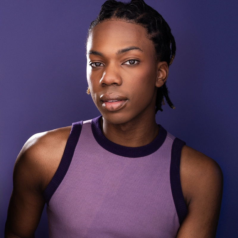 A young Black person with short dreadlocks wears a sleeveless lavender top with dark purple trim, posing against a solid purple background and looking confidently at the camera. Their expression is calm and relaxed, capturing the poised presence of Bryson Brunson.