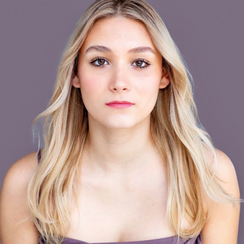 A young woman with long, straight blonde hair and light skin looks directly at the camera. She has a neutral expression, brown eyes, and wears a sleeveless, purple top. The background is a solid, muted purple color, creating an elegant setting reminiscent of an Annabelle Bryant novel.