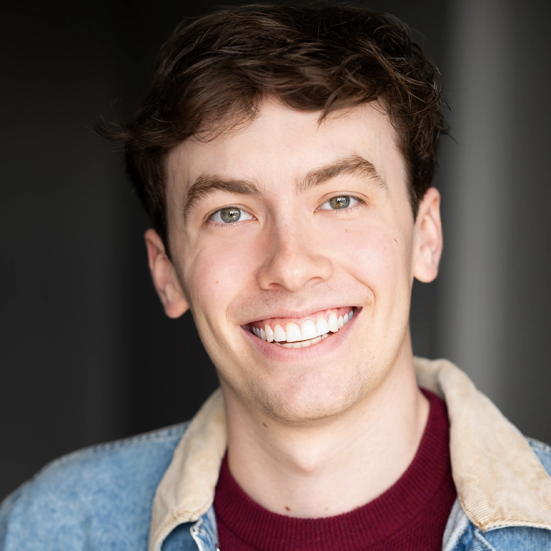 A young man with light skin, short brown hair, and green eyes smiles broadly. Trace Burchart wears a red shirt and a denim jacket with a beige collar. The background is softly blurred and dark, highlighting his face. 231 characters.
