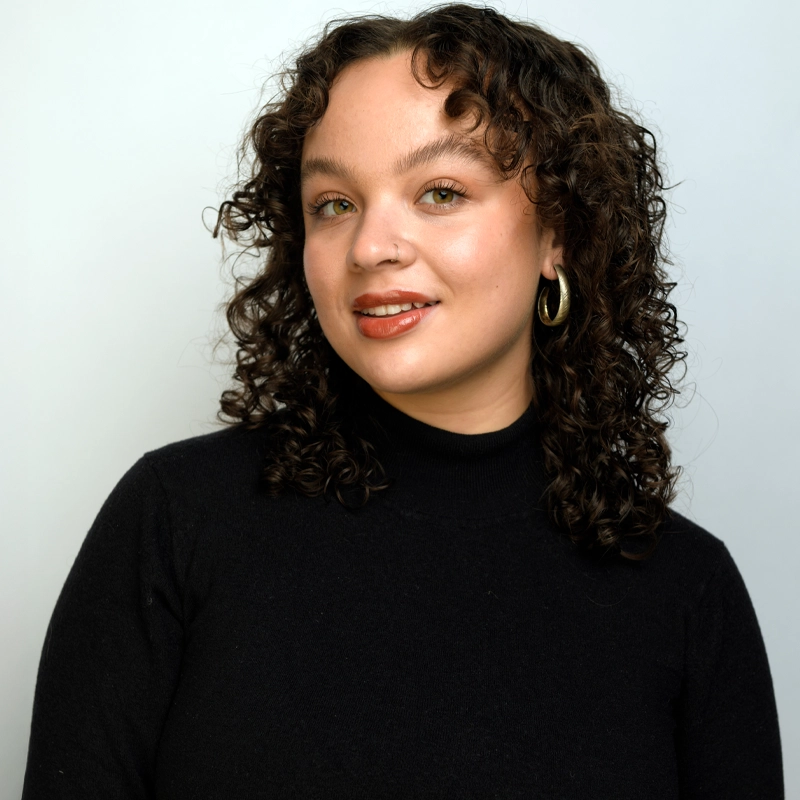 A woman with light skin and curly brown hair smiles softly. She wears gold hoop earrings, natural makeup, and a black turtleneck. The background is plain light gray. Her body is angled slightly, with her head turned toward the camera, capturing the poised confidence of Olivia Busche.