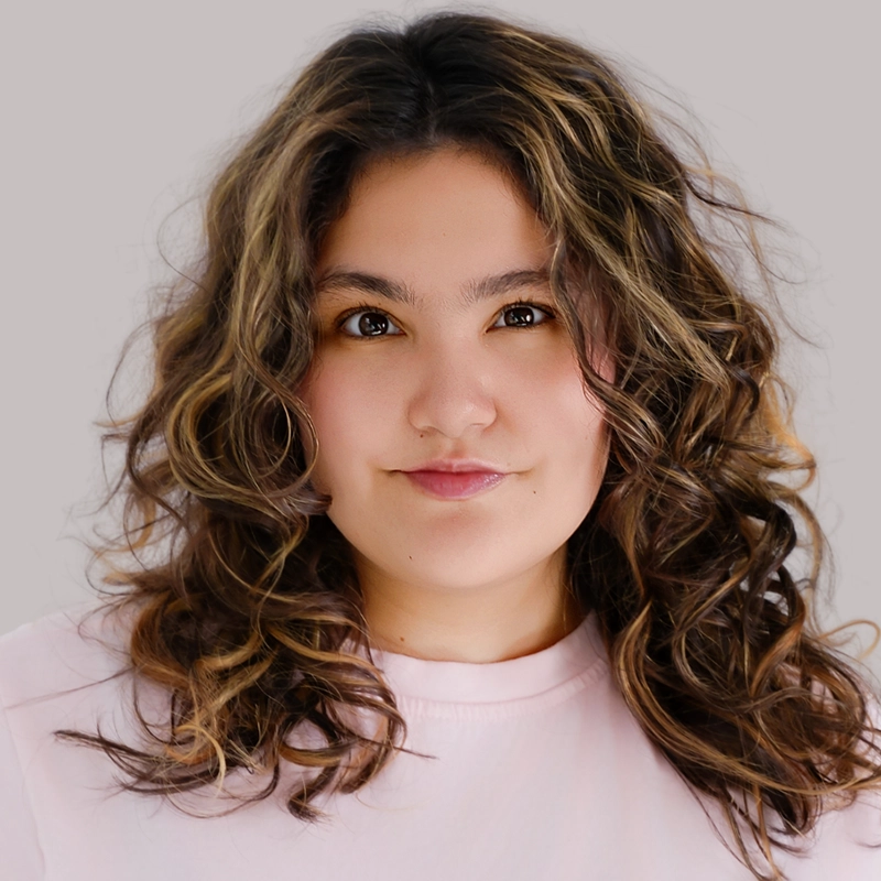 Celeste Zwingel, a person with wavy brown hair and light highlights, wears a pale pink shirt while looking directly at the camera with a slight smile. The background is plain light gray.