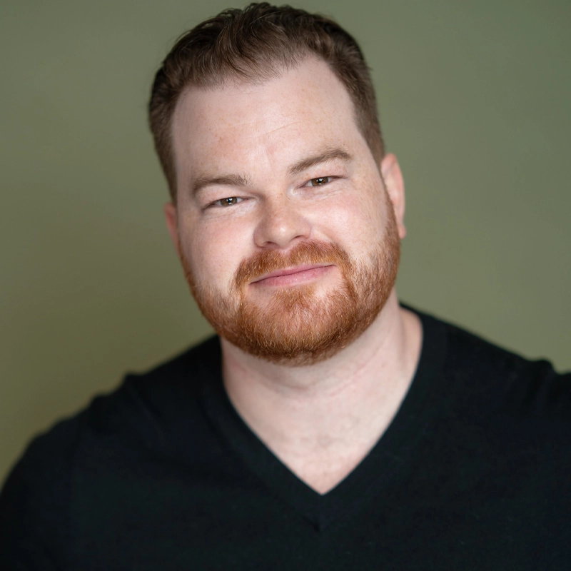 A man with fair skin, short red hair, and a full red beard smiles slightly. He wears a black V-neck shirt and is posed against a plain olive-green background. The lighting is soft and even, highlighting the subtle confidence of Oliver Callahan.