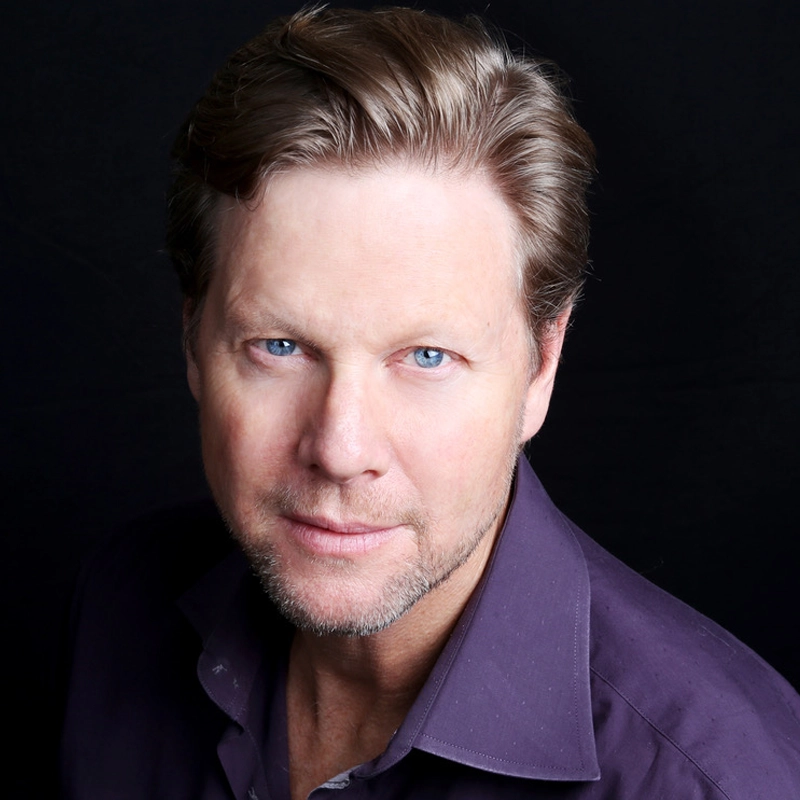 A middle-aged man with light skin, blue eyes, and short, light brown hair is wearing a purple collared shirt. Alan Campbell is looking directly at the camera with a slight smile. The background is plain and black, giving the portrait a professional feel.