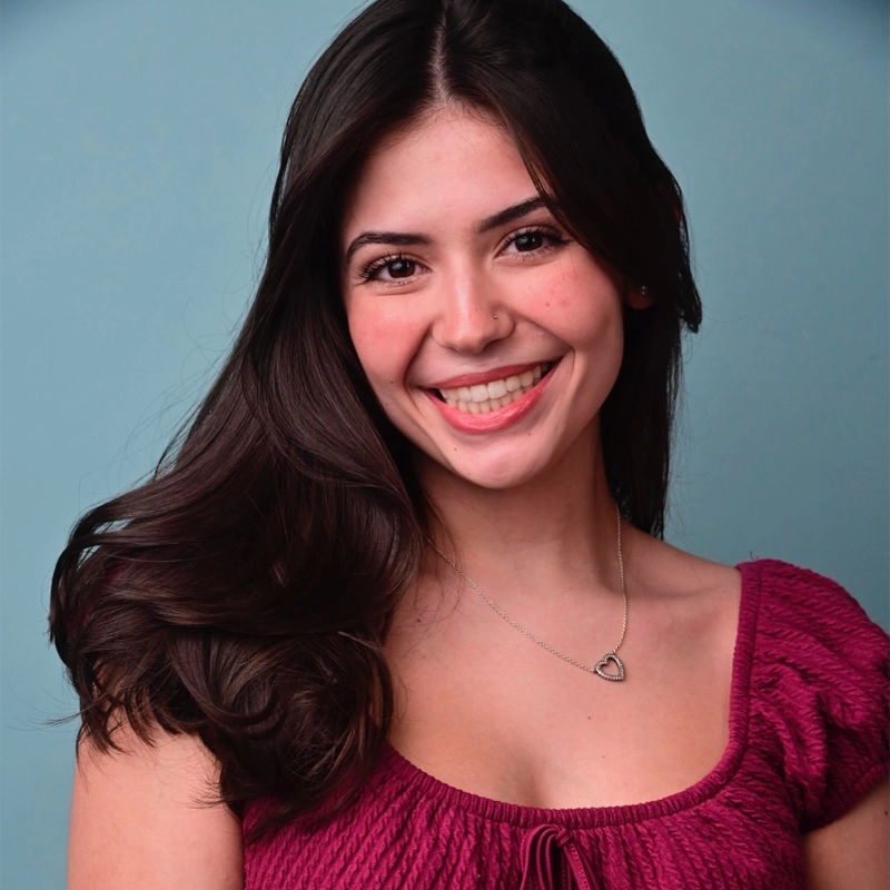 Young woman with long dark hair smiles warmly at the camera. She wears a magenta top with short sleeves and a silver heart-shaped necklace. The background is a solid light blue. Her pose is relaxed, and her makeup is natural.