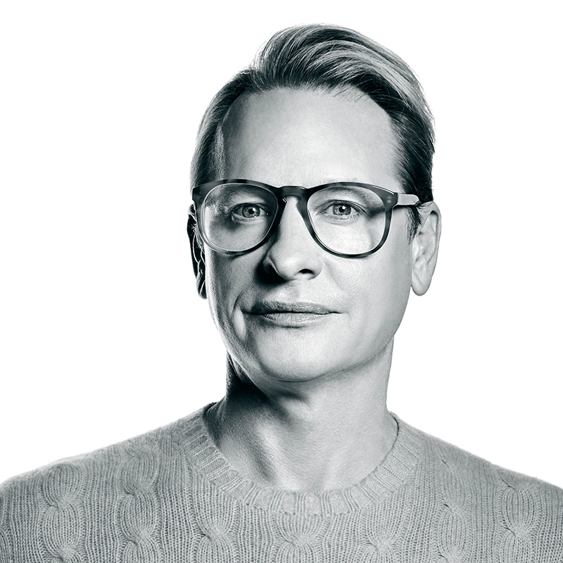 Black-and-white photo of a person with short, neatly combed hair, wearing large round glasses and a knit sweater. They face forward with a neutral expression reminiscent of Carson Kressley’s signature poise. The background is plain white, and the lighting highlights facial features clearly.