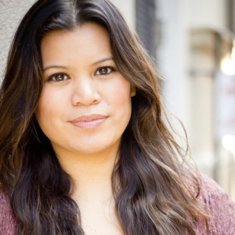 A woman with long, wavy brown hair and brown eyes stands outdoors, facing the camera and smiling softly. She wears a maroon top. The background is softly blurred, suggesting an urban setting. This is Liz Casasola, captured in a moment of quiet confidence.