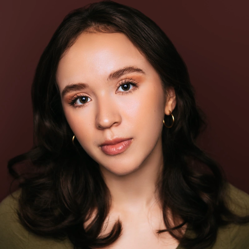 A young woman with wavy brown hair and blue eyes looks at the camera. She has light skin, wears gold hoop earrings, subtle makeup, and a green top. The background is a solid deep burgundy color. With a neutral, calm expression, Celeste Zwingel exudes quiet confidence in this portrait.