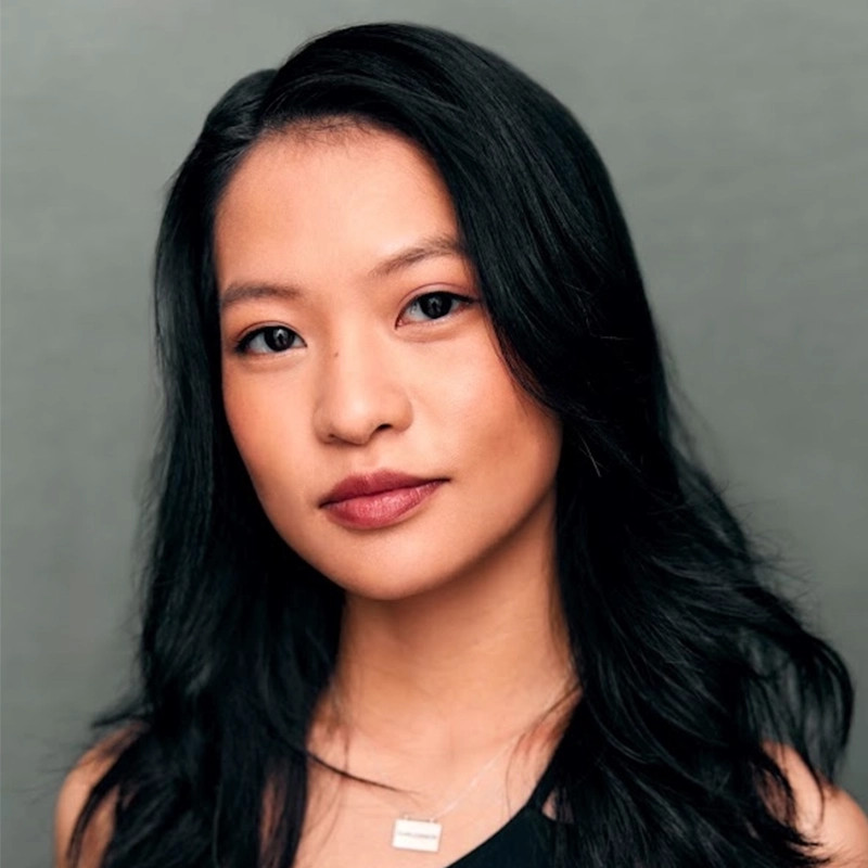 A young woman with long, wavy black hair and light makeup looks at the camera. She wears a black top and a silver necklace with a rectangular pendant. The background is plain and gray. The lighting is soft and even, drawing gentle attention to her features in a style reminiscent of Kendall Tubbs.