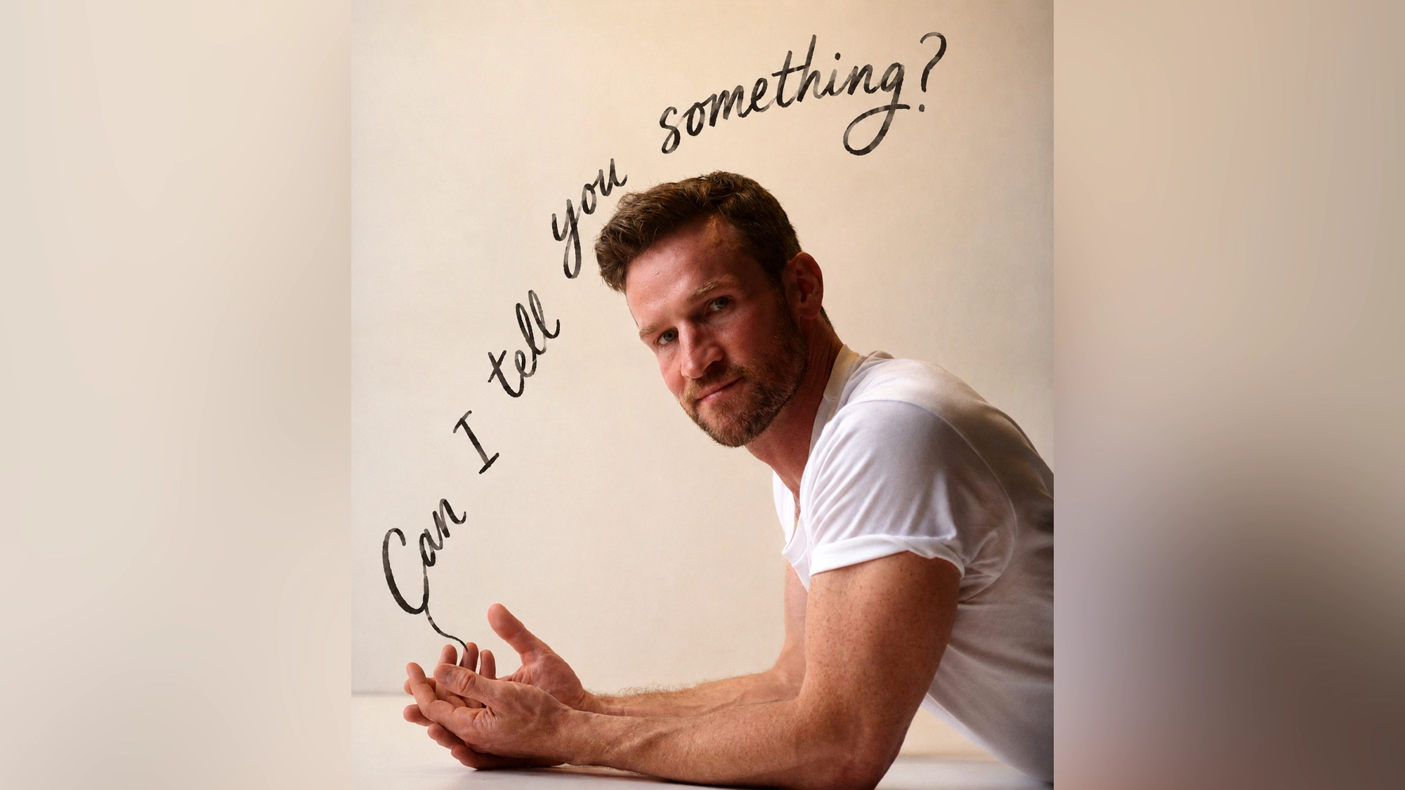 A man in a white t-shirt leans on a surface, looking at the camera. Black handwritten text arcs above him, reading “Can I Tell You Something?” The beige, plain background keeps the focus on Claybourne Elder and his inviting expression.