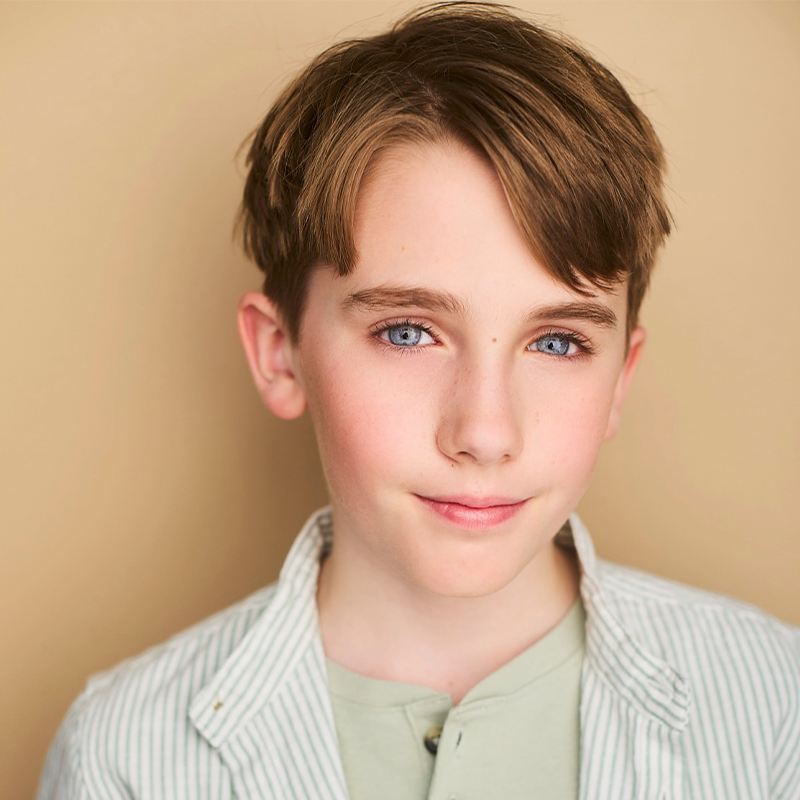 A young boy with light skin, blue eyes, and short brown hair smiles softly. He wears a light green shirt and a white-striped button-up shirt. The background is a plain beige color, and the lighting is soft and even.