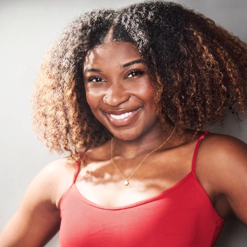 Grace Conrad, a young woman with curly, shoulder-length hair, smiles warmly. She wears a red camisole and a delicate gold necklace. She stands against a plain gray background, with sunlight casting soft shadows on her face and shoulders.