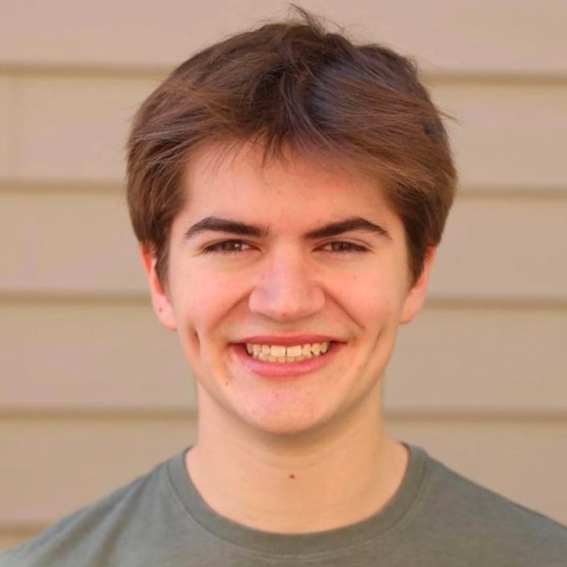 Cooper Brown, a young person with short brown hair and dimples, smiles at the camera. They are wearing a green shirt and standing in front of a tan, horizontal-paneled wall. The lighting is bright and even, giving the scene a cheerful appearance.