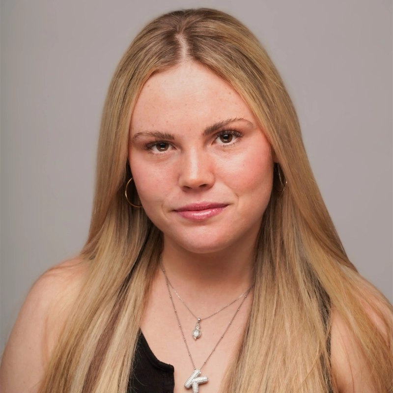 A young woman with straight, long blonde hair and fair skin smiles softly. She wears hoop earrings, layered silver necklaces, a black top, and has natural makeup. The background is a plain light gray.