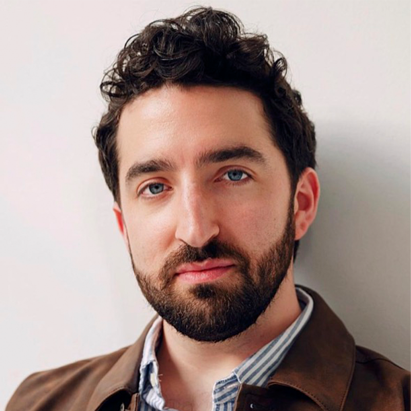 A man with wavy dark hair, a trimmed beard, and blue eyes faces the camera. He wears a brown jacket over a light blue striped shirt. The background is plain and light-colored, and he has a neutral expression on his face.