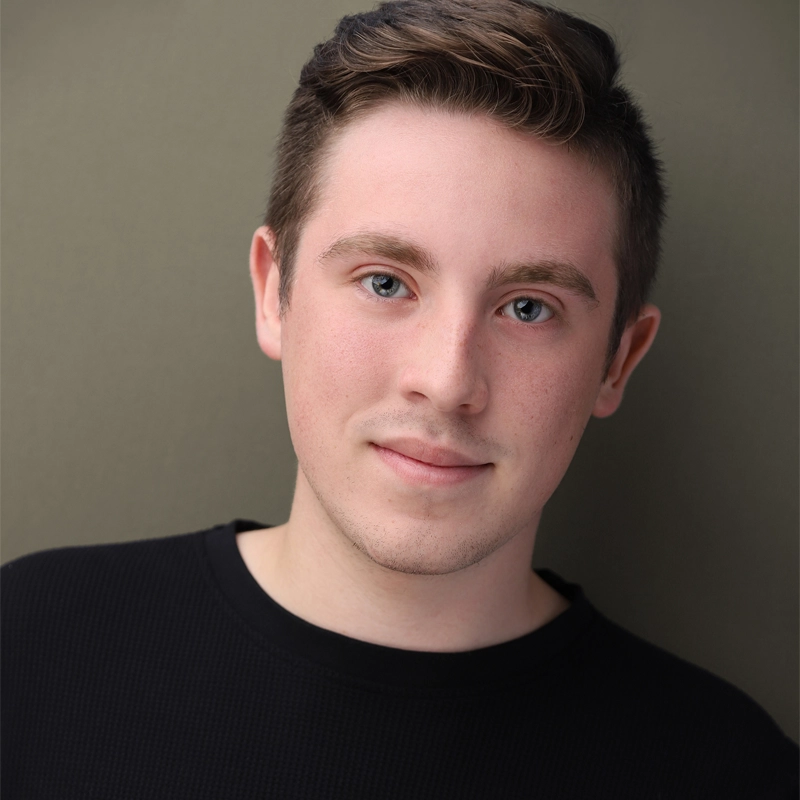 A young man with short brown hair and blue eyes wears a black sweater, smiling softly at the camera. Aaron Crowell poses against a plain, olive-green background, lit with soft, even lighting that highlights his clear skin and relaxed expression.