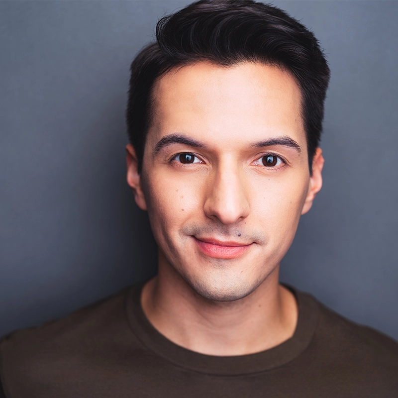 A young man with short dark hair and fair skin looks directly at the camera, smiling slightly. He wears a dark-colored crew neck shirt. The background is a plain gray color, creating a simple and clean portrait—a style reminiscent of Fabi Aguirre’s understated yet captivating presence.