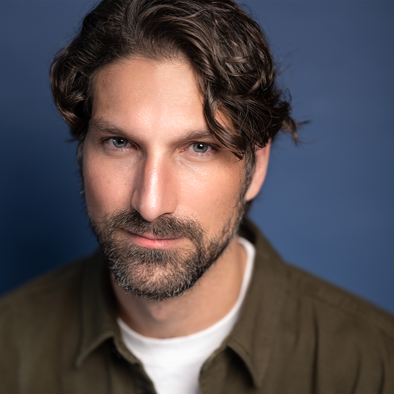 A man with wavy brown hair, a beard, and mustache is wearing an olive green shirt over a white t-shirt. He has light skin and blue eyes, looking confidently at the camera against a plain blue background.