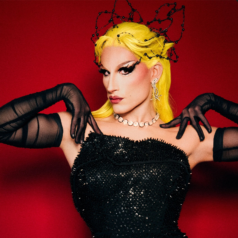 A person with bright yellow hair and dramatic black eyeliner poses against a red background. They wear a beaded crown, sparkly earrings, a jeweled necklace, a black beaded strapless dress, and sheer black gloves, with hands elegantly raised near their shoulders.