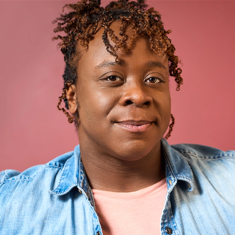 A person with shoulder-length, curly brown hair wears a light blue denim shirt over a pink t-shirt. They are looking toward the camera against a solid red background.