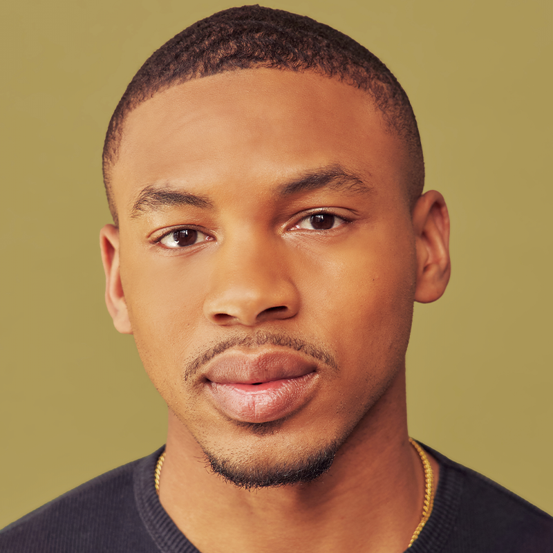 A young man with short dark hair and trimmed facial hair faces forward, wearing a black shirt and gold chain. The background is a solid light olive color. His expression is neutral and direct.